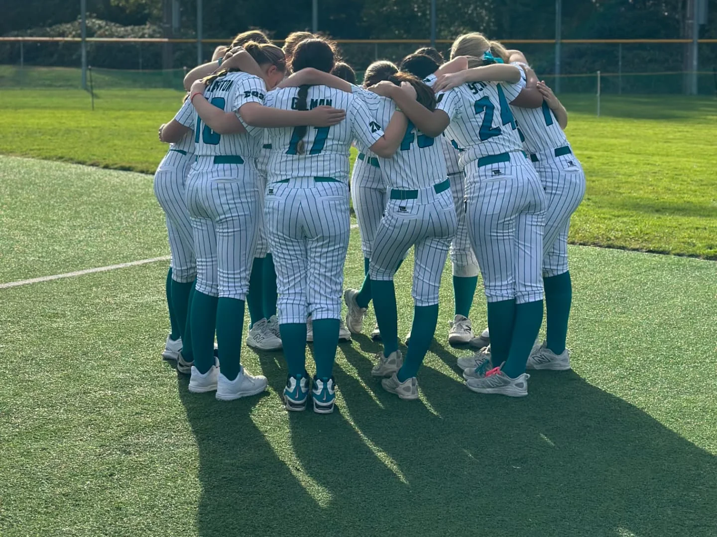 United Softball players in white pinstripe uniforms with teal accents gathered in a team huddle on the field.